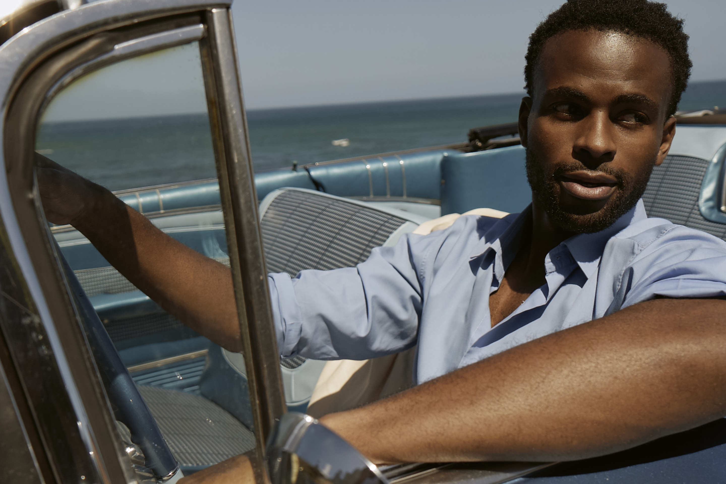Lifestyle campaign portrait of a man in a blue shirt seated in a vintage convertible by the sea.